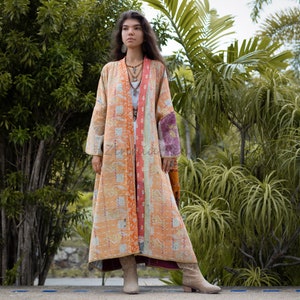 May include: A long, orange and pink quilted jacket with a floral pattern. The jacket has a long, flowing silhouette and is worn by a person standing in front of a green leafy background.