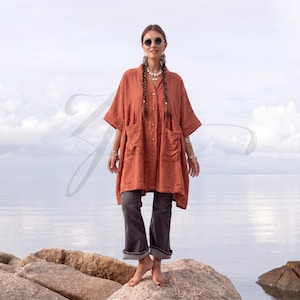 May include: A rust-colored, button-down tunic with short sleeves, a V-neckline, and two large front patch pockets. The garment is paired with dark gray, straight-leg trousers that have a cuffed hem, creating a layered, casual ensemble.