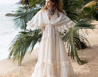 May include: A white maxi dress with a flowing, tiered skirt and a V-neckline. The dress has a cape-like top with ruffles and is made of a lightweight fabric. The model is wearing the dress on a beach with palm trees in the background.