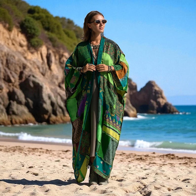 Full-length front view of the Harper Jade Kimono in Emerald Oasis, styled on a sandy beach with rocky cliffs and turquoise waves in the background. The patchwork robe, made from upcycled vintage silk saris, flows gracefully in jewel-toned emerald, teal, saffron, and amethyst hues. A bohemian duster designed for festivals, travel, and resort wear.