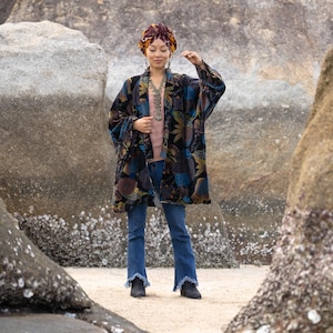 Op de afbeelding: Een vrouw draagt een lange, open kimono van fluweel met een bloemenpatroon in blauw, bruin en goud. Ze draagt ook een tulband, een roze top, een blauwe jeans en zwarte laarzen. De kimono is het centrale punt van de afbeelding.