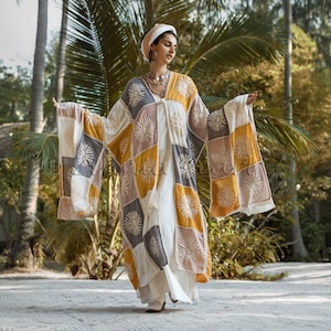 Puede incluir: Una mujer lleva un kimono de patchwork blanco, amarillo y gris con detalles florales bordados. El kimono tiene una silueta suelta y fluida y se lleva sobre un vestido blanco. La mujer está de pie en un entorno tropical con palmeras en el fondo.