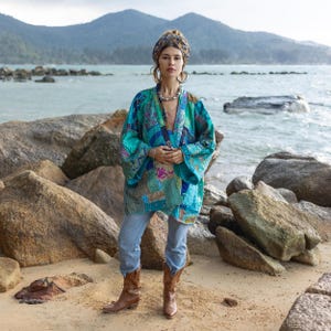 May include: A woman wearing a blue and green patchwork kimono with a brown head scarf and brown cowboy boots. She is standing on a beach with rocks in the background.