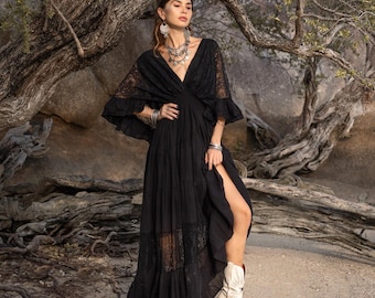 May include: A black maxi dress with a plunging neckline and lace detailing. The dress features flared sleeves and a high slit, paired with white cowboy boots. The model is wearing silver jewelry and a headpiece, posing outdoors near a tree.