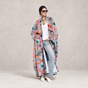 Könnte beinhalten: Ein langer, vorne offener Kimono-Mantel mit hellblauem Grund und einem lebendigen Blumenmuster in Orange-, Rot- und Grüntönen. Das Kleidungsstück wird über einem schlichten weißen Tanktop und einer hellblauen Jeans getragen, ergänzt durch ein schwarzes Kopftuch.