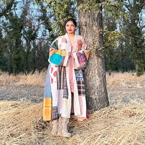 Puede incluir: Una bata tipo kimono patchwork en una variedad de colores y patrones, incluyendo telas florales, geométricas y lisas. La bata es larga, con mangas anchas y un ajuste relajado. El fondo es un campo con un árbol.