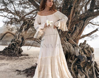 Puede incluir: Vestido largo color crema con hombros descubiertos, mangas largas y puños con volantes. El vestido presenta una falda escalonada y está hecho de una tela texturizada. La imagen incluye el texto "ZINNIA PE".