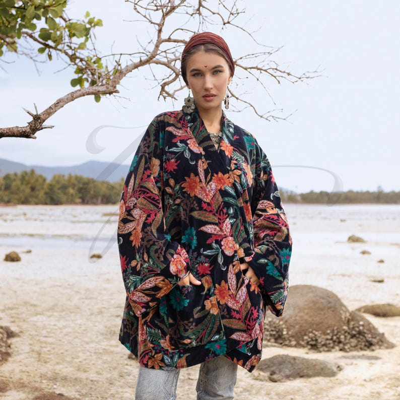 Close-up of the Mei Velvet Kimono Duster in Nocturne Flora by ZinniaPearl. The model stands near the shoreline, framed by a graceful tree branch, her gaze calm and confident. The velvet fabric glows with hand-embroidered florals in jewel hues of magenta, ochre, and jade. A portrait of handcrafted beauty, feminine power, and timeless bohemian style.