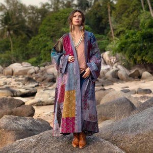 May include: A long, patchwork kimono-style coat in shades of purple, pink, blue, and gold. The coat features a variety of patterned fabric squares sewn together. The person is wearing a peach-colored top, blue jeans, and brown boots. The background is a beach scene.