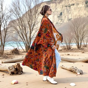 Side view of the Amber Flora Harper Jade Kimono Robe, flowing dramatically as the model stands on a coastal beach with driftwood and bare trees. This floor-length plus size velvet duster coat is crafted from soft cotton velvet in warm amber tones with intricate floral motifs. The oversized fit and deep pockets offer boho elegance and comfort, perfect for autumn walks, holiday celebrations, or layered festival styling.