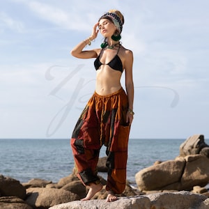 May include: A woman wearing a black bikini top and colorful patchwork pants stands on a rock by the ocean. She is wearing a headband and earrings.
