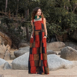 May include: A long, red, black, and brown patchwork dress with a square neckline and thin straps. The dress has pockets on the sides and is worn by a person with long brown hair and green earrings.