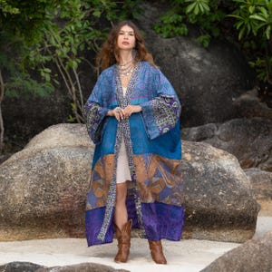 May include: A long, flowing kimono-style robe in shades of blue, purple, and brown. The robe features a patchwork design with patterned sleeves and trim. The model is wearing brown cowboy boots.