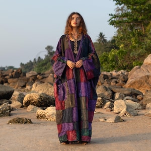 May include: A patchwork robe in shades of purple, red, and green. The long, flowing garment features a geometric pattern and two large pockets. The robe is worn on a sandy beach with rocks and trees in the background.