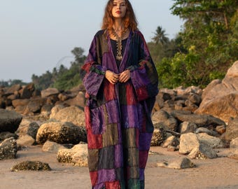 May include: A patchwork robe in shades of purple, red, and green. The long, flowing garment features a geometric pattern and two large pockets. The robe is worn on a sandy beach with rocks and trees in the background.
