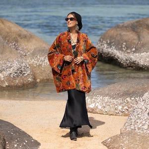 May include: An orange velvet kimono with a floral pattern, worn over a black dress. The kimono features a vibrant mix of colors, including red, green, and blue. The outfit is accessorized with jewelry and sunglasses.
