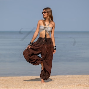 Könnte beinhalten: Eine Person trägt ein silbernes Bustier und eine braune Haremshose an einem Sandstrand. Die Hose hat ein Patchwork-Design und ist an den Knöcheln gerafft. Sie trägt eine Sonnenbrille, eine Halskette und Armbänder.