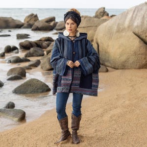 May include: A woman wearing a blue and black patterned hooded jacket with a geometric design. She is standing on a sandy beach with large rocks in the background.