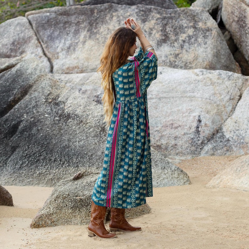 3. Side-back view of the Cora Midi Dress in Peacock Rose photographed outdoors on a rocky beach setting. The relaxed oversized silhouette showcases flowing cambric cotton fabric with teal artisan print, magenta contrast side panels, and softly draped kimono sleeves. Styled with brown leather boots, the dress captures movement, comfort, and elevated bohemian ease.