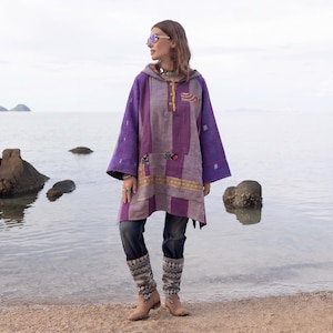May include: A hooded tunic featuring a patchwork design in shades of purple and grey, with a buttoned placket, long sleeves, and decorative embroidered accents. The garment is paired with dark grey denim jeans and patterned leg warmers.