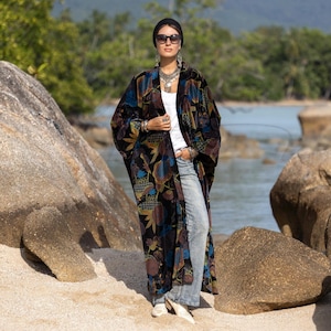 Pode incluir: Um longo quimono de veludo preto com um padrão floral em tons de azul, castanho e dourado. O quimono é usado sobre uma blusa branca e jeans claros. O conjunto é complementado com joias e óculos de sol.