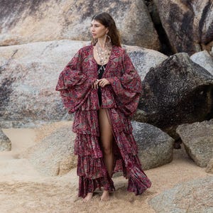 May include: A long, flowing burgundy kimono with a paisley pattern. The kimono features tiered ruffles and wide sleeves. The garment is open in the front and worn on a sandy beach with large rocks in the background.