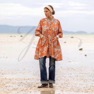 May include: An orange button-down tunic dress with a floral pattern, featuring three-quarter sleeves and pockets. The model is wearing blue jeans and black boots. The background is a beach with a cloudy sky.