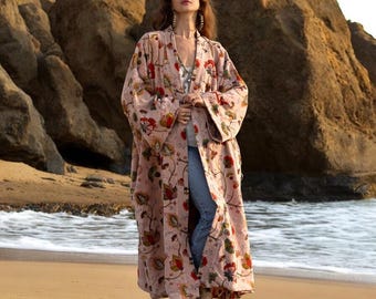 May include: A long, flowing, light pink kimono-style robe with a floral pattern in red, green, and yellow. The robe is open in the front and worn over a white top and blue jeans. The model is wearing white boots and standing on a sandy beach.