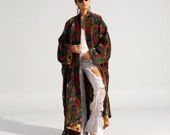Puede incluir: Un abrigo largo de terciopelo verde oscuro con un estampado floral vibrante en tonos rojos, amarillos y azules. Se lleva sobre una camiseta blanca sin mangas y unos vaqueros blancos acampanados y desgastados con bajos deshilachados y botas.