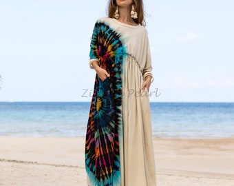 May include: A long, flowing dress with a tie-dye pattern on one side and a solid beige color on the other. The dress has a cinched waist and a relaxed fit. The model is standing on a sandy beach with the ocean in the background.