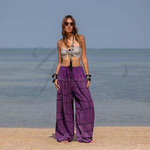 May include: Purple wide-leg pants with a grid-like pattern. The pants have a drawstring waist and are paired with a gray tube top. The model is wearing sunglasses and jewelry, standing on a sandy beach with the ocean in the background.