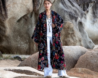 May include: A long, dark floral patterned robe with red, blue, and green accents. The robe is worn over a white top and light blue pants. The robe's design features a variety of flowers and leaves.