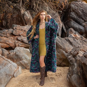May include: A woman in a long, patterned robe with green, purple, and white floral designs. She wears a gold dress and brown boots. The background shows rocks and sand, creating a natural setting.
