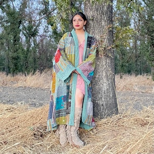 May include: A long, patchwork quilt jacket in shades of blue, green, yellow, and red. The jacket has a shawl collar and long sleeves. The person wearing the jacket is standing in a field with tall grass.