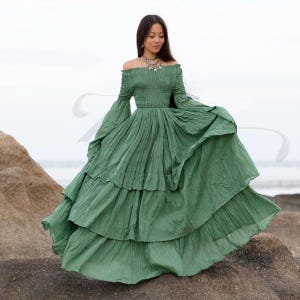 Lucia by ZinniaPearl is a plus-size-friendly boho dress in raw cotton, handcrafted in an earthy Elderfern Green shade. With a flowing tiered skirt, stretch smocking, and long flared sleeves, this gown is made for moonlit weddings, forest rituals, or beach elopements. Its off-shoulder neckline and breathable fabric make it a dreamy fit for brides, goddesses, and maternity moments in nature.