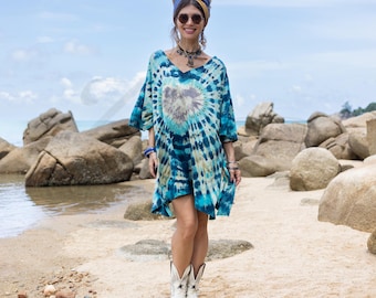 May include: A blue and turquoise tie-dye tunic dress with a V-neck and short sleeves. The dress features a heart-shaped design in the center. The person is wearing white cowboy boots, sunglasses, and a blue headband. The background is a beach with rocks and the ocean.