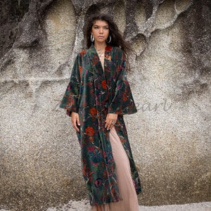 May include: A long, green velvet robe with a floral print. The robe has a shawl collar and long sleeves. The model is wearing a nude slip dress underneath the robe.