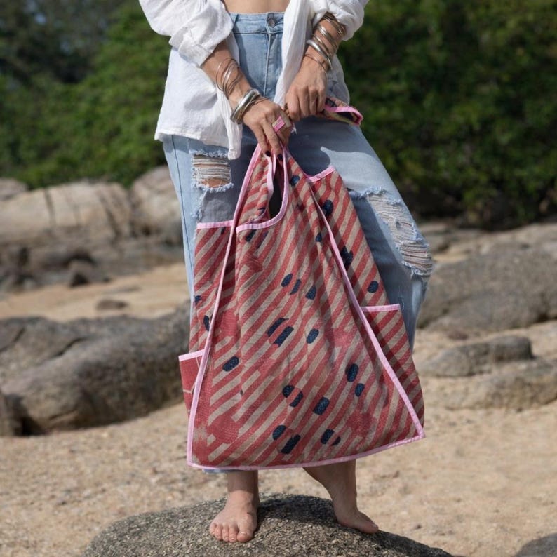Model holding the Kaia Dusty Rose Polka Kantha hobo bag, a reversible vintage quilt shoulder tote handcrafted from upcycled cotton sari textiles. The soft dusty rose base features diagonal stripe texture with polka-inspired detailing and hand-stitched Kantha running stitch, styled casually with denim against a rocky outdoor backdrop.