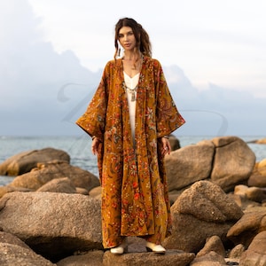May include: A long, mustard-colored kimono with a floral pattern in red, orange, and white. The kimono is worn over a white dress and paired with white shoes. The background features a rocky beach and the ocean.
