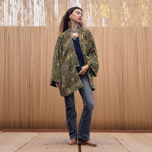 Model wearing an emerald green and gold hand block-printed velvet kimono jacket with wide sleeves, styled casually with jeans and a navy top, standing against a natural bamboo wall background.