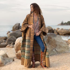 May include: A long, brown patterned kimono with a green and black geometric design. The kimono is worn over a light pink top and blue jeans. The person is standing on a beach with rocks in the background.