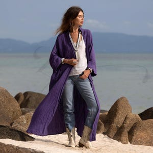 May include: A long, flowing, purple duster coat. The coat is worn over a white top and blue jeans. The person is wearing white cowboy boots and silver jewelry. The background is a beach with rocks and the ocean.
