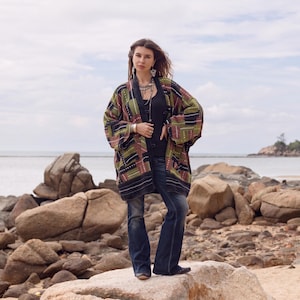 May include: A long-sleeved, open-front kimono jacket featuring a patchwork design in shades of olive green, dark brown, and black with white stitched detailing. The garment is worn over a black tank top and dark blue flared denim jeans.