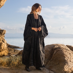 May include: A woman wearing a long, black, flowing robe with a textured pattern. She is standing on a rocky beach with the sea in the background.