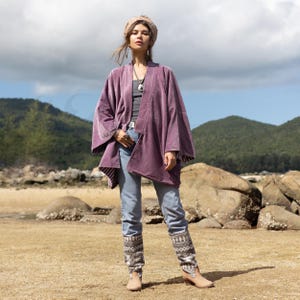 May include: A woman wearing a purple velvet kimono with a light blue denim shirt and jeans. She is wearing brown knitted leg warmers and brown boots. The woman is standing on a beach with rocks and sand in the background.
