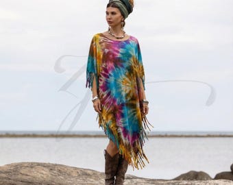 May include: A colorful tie-dye poncho with fringe trim. The poncho features a spiral pattern in shades of blue, pink, yellow, and orange. The model is wearing brown boots and a matching head wrap. The background is a seascape.