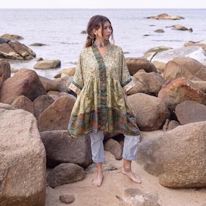 May include: A long-sleeved, button-down tunic featuring a multi-colored pattern with shades of beige, green, and blue. The garment has a V-neckline, two front pockets, and a flared silhouette, paired with light blue denim jeans rolled at the ankles.