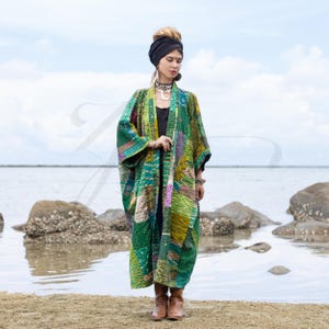May include: A woman wearing a long, green, patchwork kimono with a black head scarf and silver necklace. She is standing on a beach with rocks and water in the background.