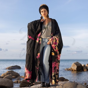Könnte beinhalten: Ein langer schwarzer Kimono mit rosa und gelben Batik-Mustern. Der Kimono hat lange Ärmel und wird über einem grauen T-Shirt und einer blauen Jeans getragen.