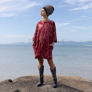 May include: A woman wearing a red tie-dye dress with a white headband and black cowboy boots. She is standing on a rock with the ocean in the background.